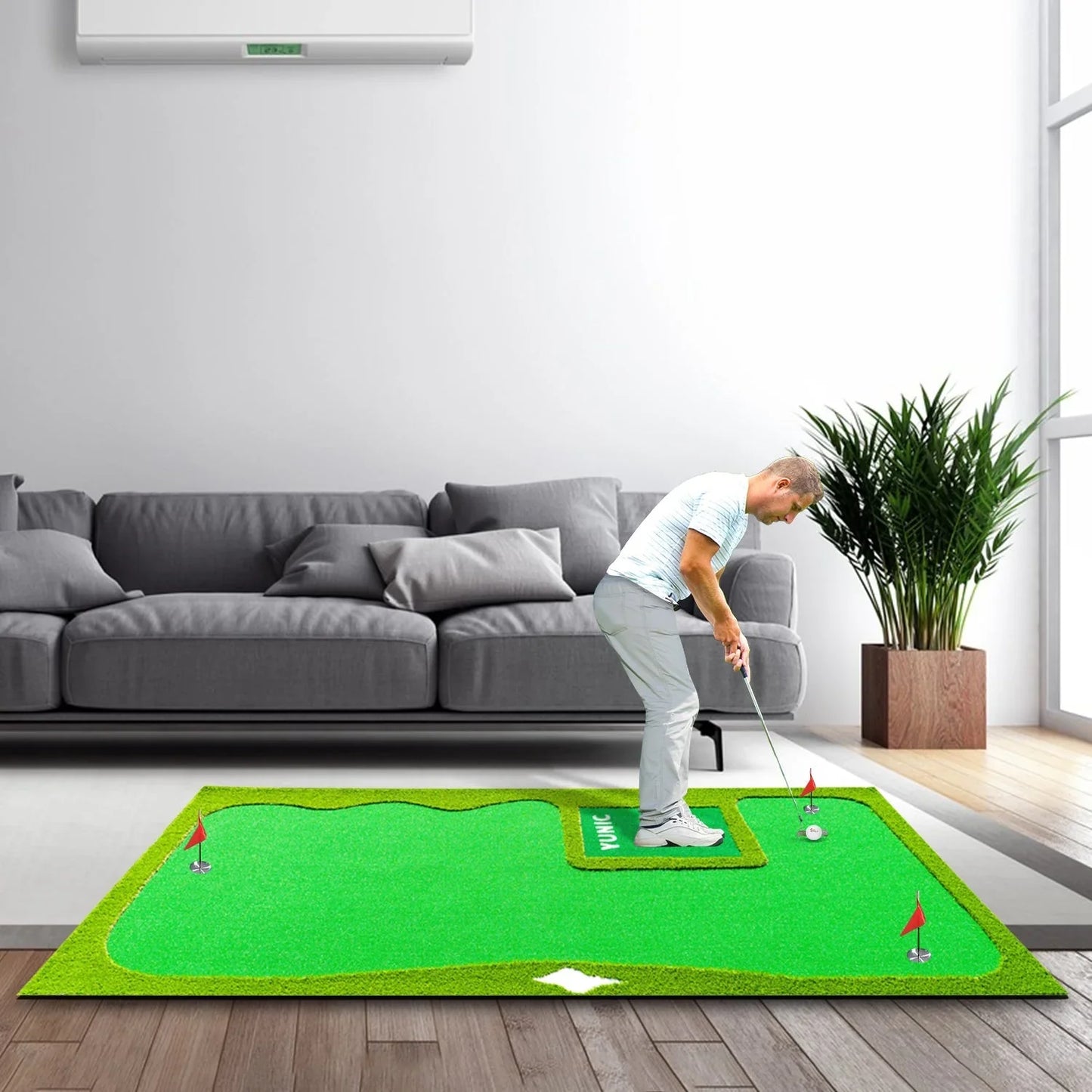Man practicing indoor golf putting on an 8x16 ft green mat near gray sofa and plant
