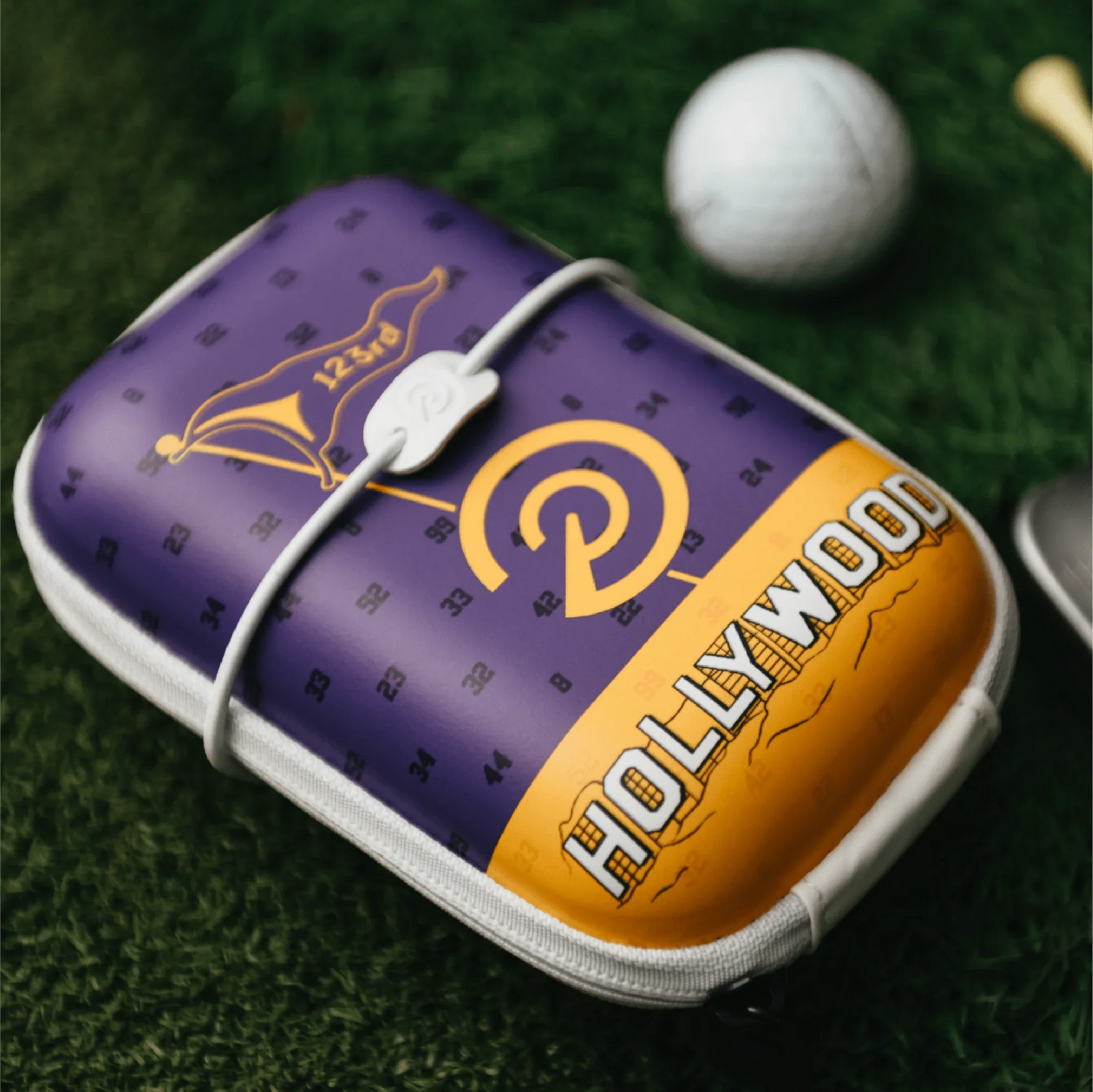 Purple and orange hard shell rangefinder case with Hollywood text on green grass next to a golf ball