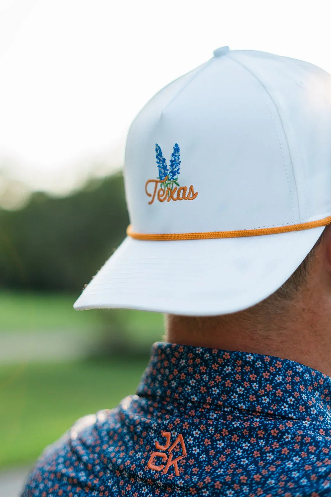 White cap with Texas bluebonnet embroidery and orange trim worn with floral patterned shirt outdoors