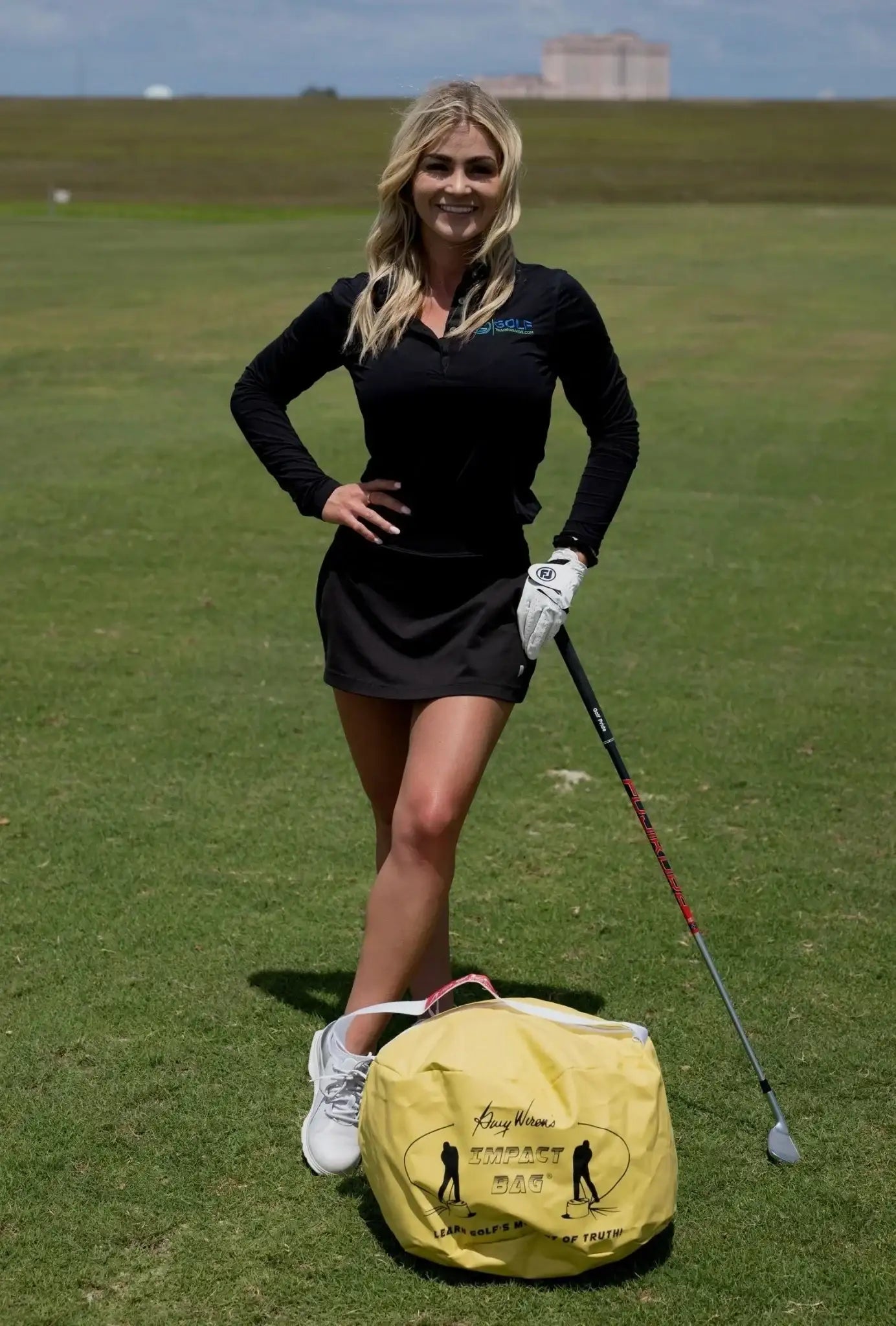 Female golfer in black outfit posing on grass with golf club and yellow Gary Wiren golf impact bag