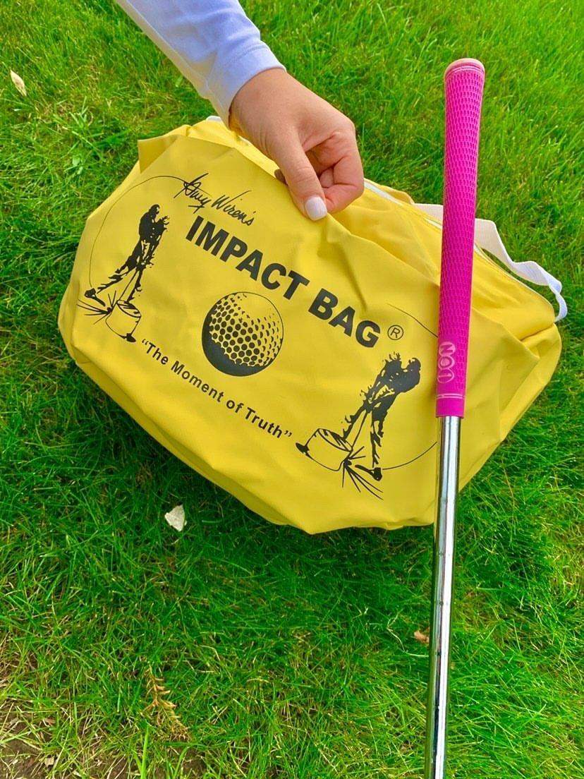 Hand holding a yellow golf Impact Bag on green grass next to a golf club with a pink grip