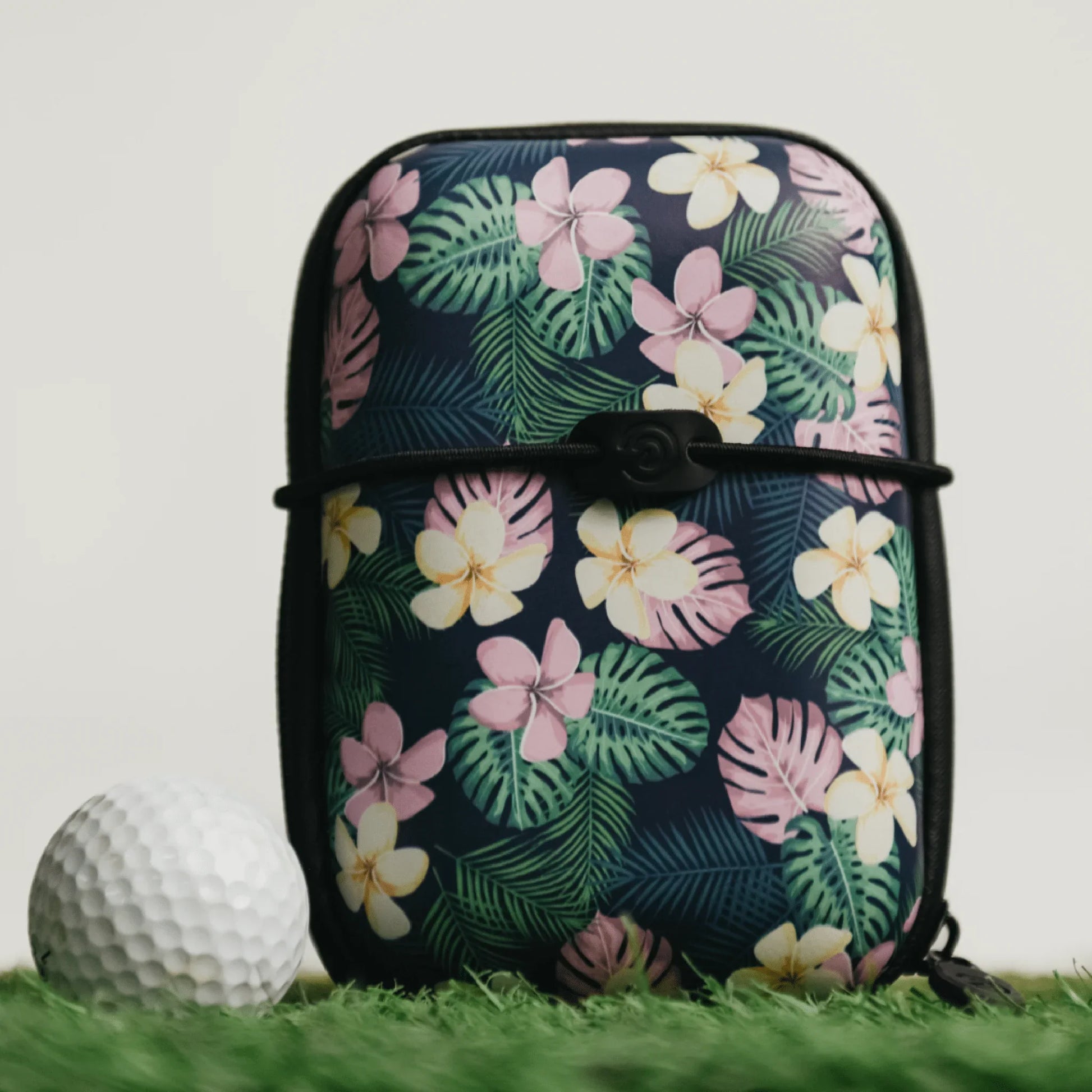 Floral hard shell rangefinder case with pink and yellow flowers beside a golf ball on green grass