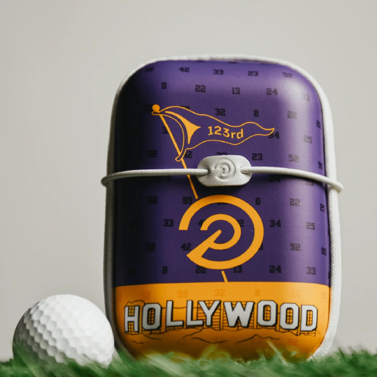 Hard shell golf rangefinder case with purple and yellow Hollywood design next to a golf ball on grass