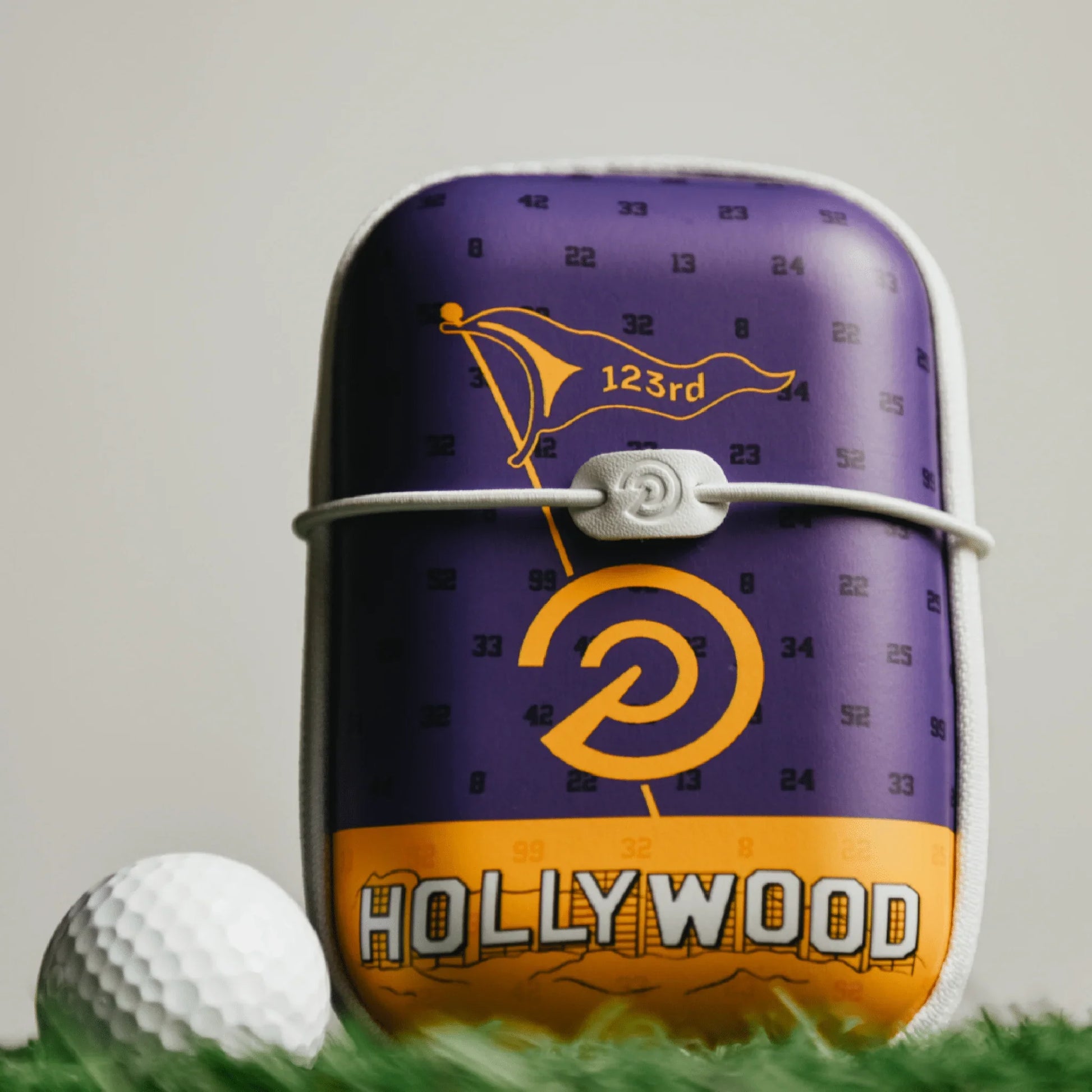 Hard shell golf rangefinder case with purple and yellow Hollywood design next to a golf ball on grass