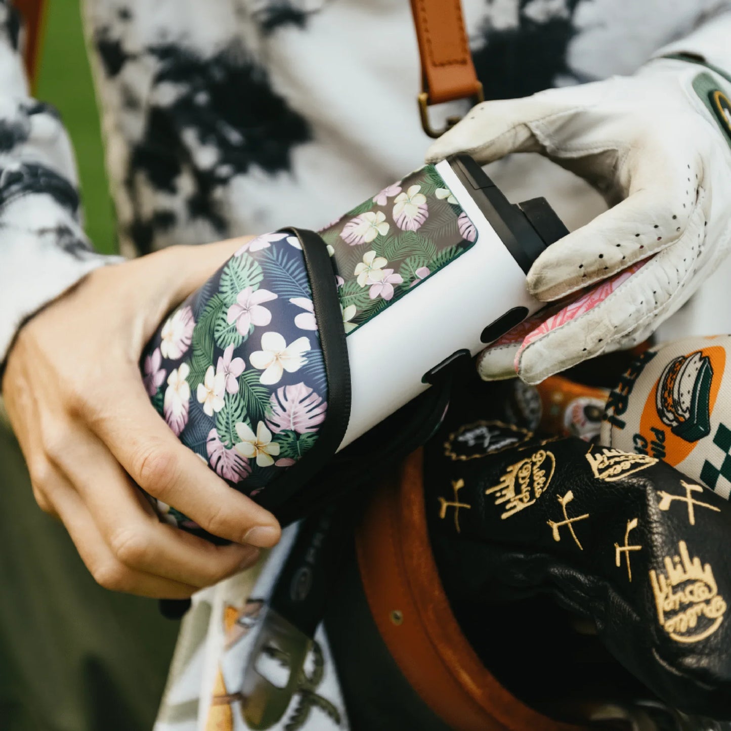 Person placing a white golf rangefinder with floral hard shell case into a golf bag with gloves and covers