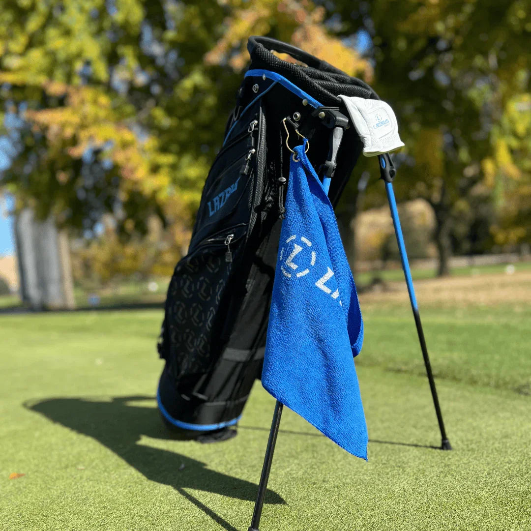 Black and blue Lazrus golf bag with stand on green grass, blue golf towel hanging, trees in background