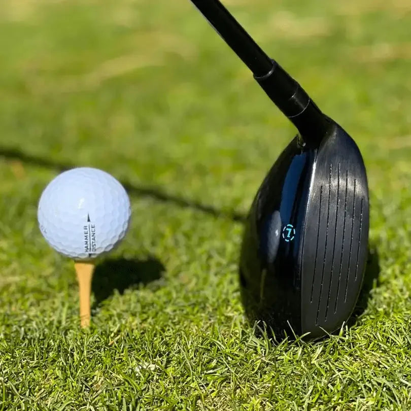 Close-up of black golf driver club face next to white golf ball on wooden tee on grassy course