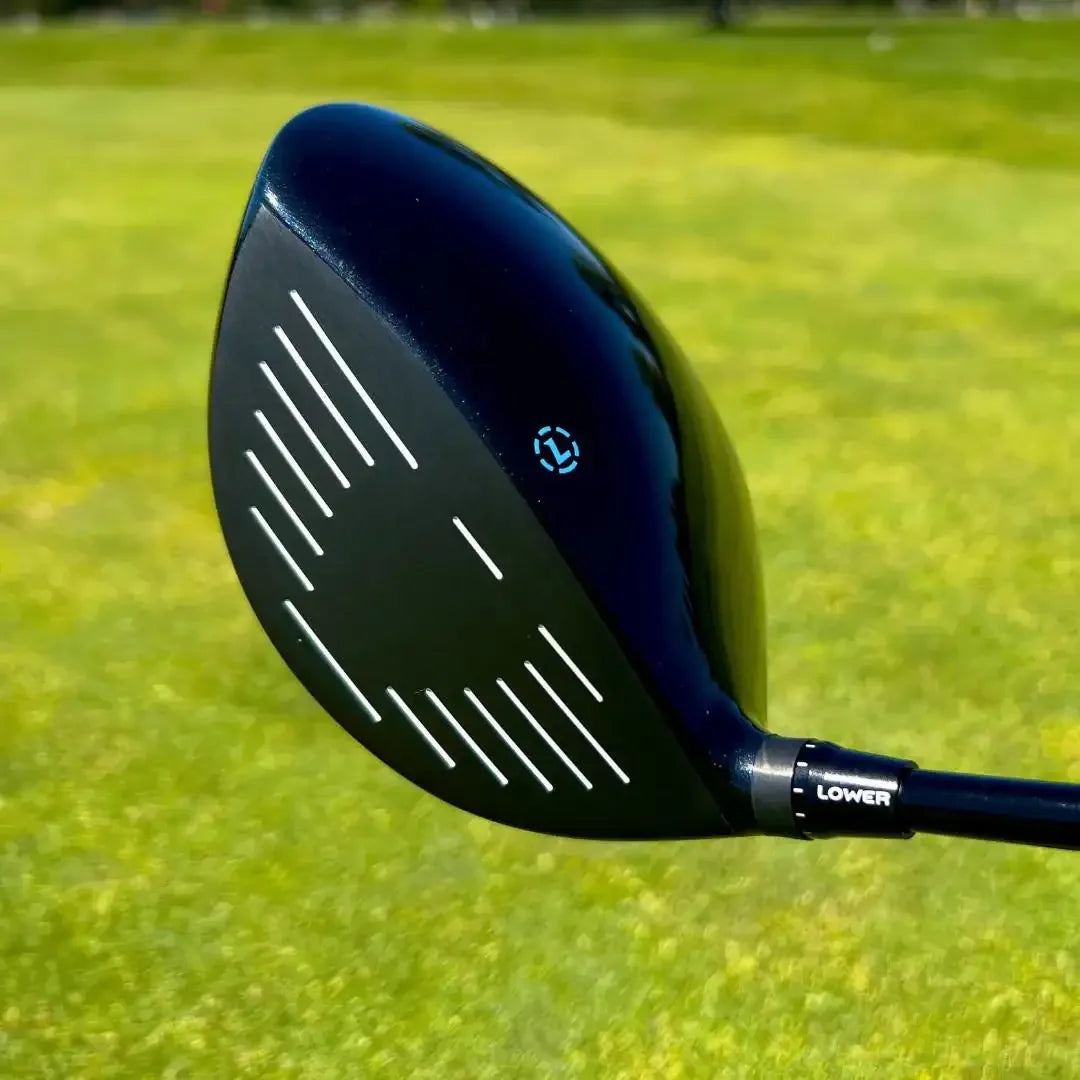 Close-up of a black golf driver club head with white grooves and adjustable shaft labeled 'LOWER' on a green grass background