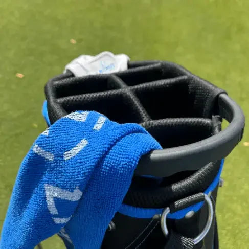 Black golf bag with multiple club slots on green grass, featuring a blue towel draped over the side