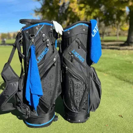 Two black Lazrus premium golf bags with blue accents and towels on a sunny golf course