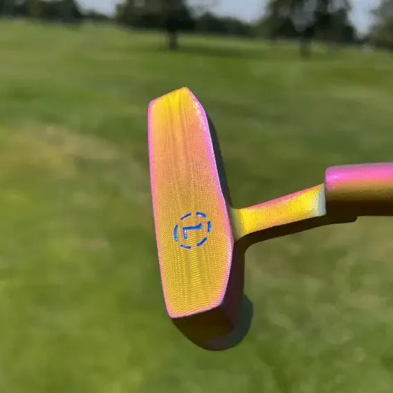 Close-up of a rainbow-colored golf putter head with milled face and blue logo on a green golf course