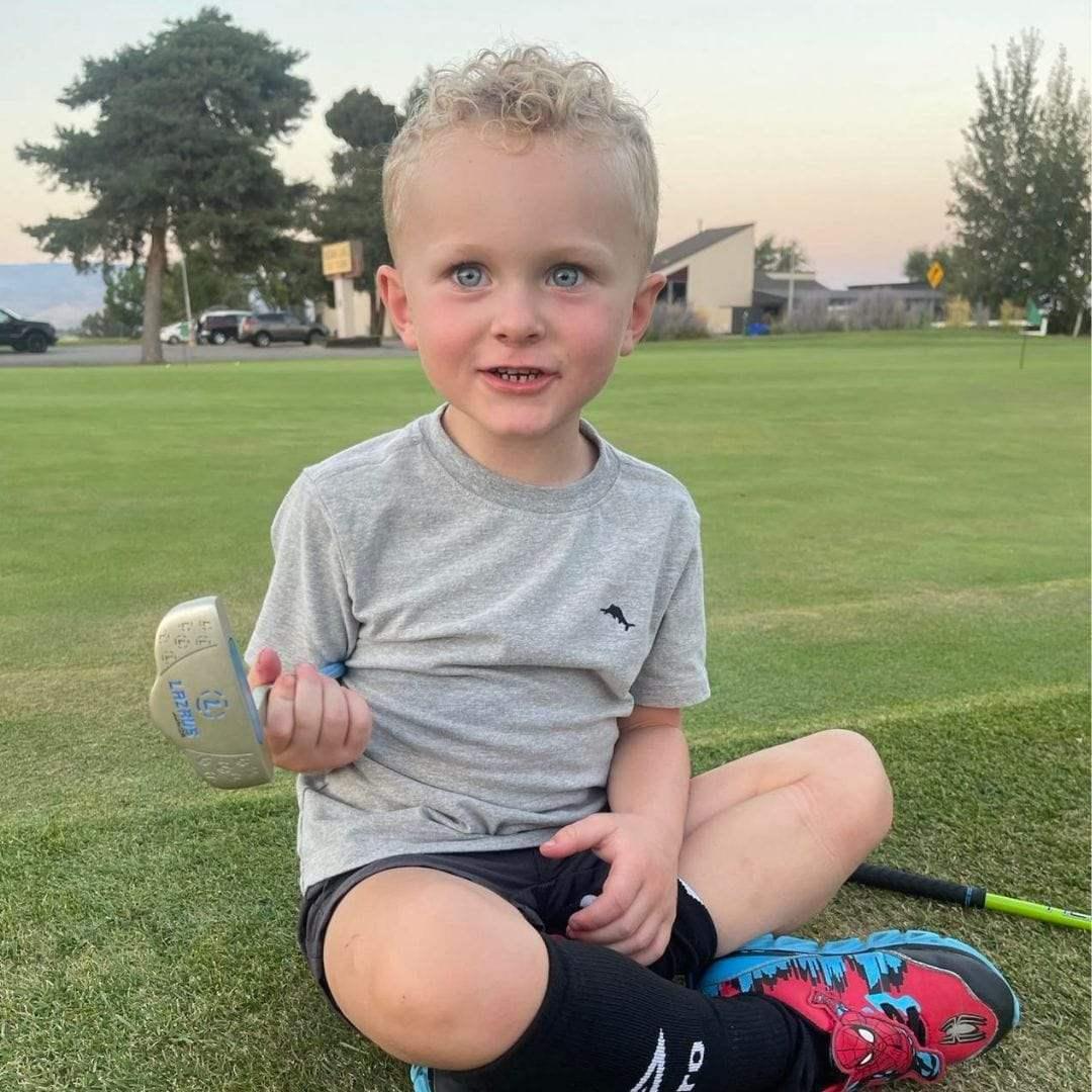 Young boy sitting on grass at golf course holding a junior golf club with trees and buildings in background