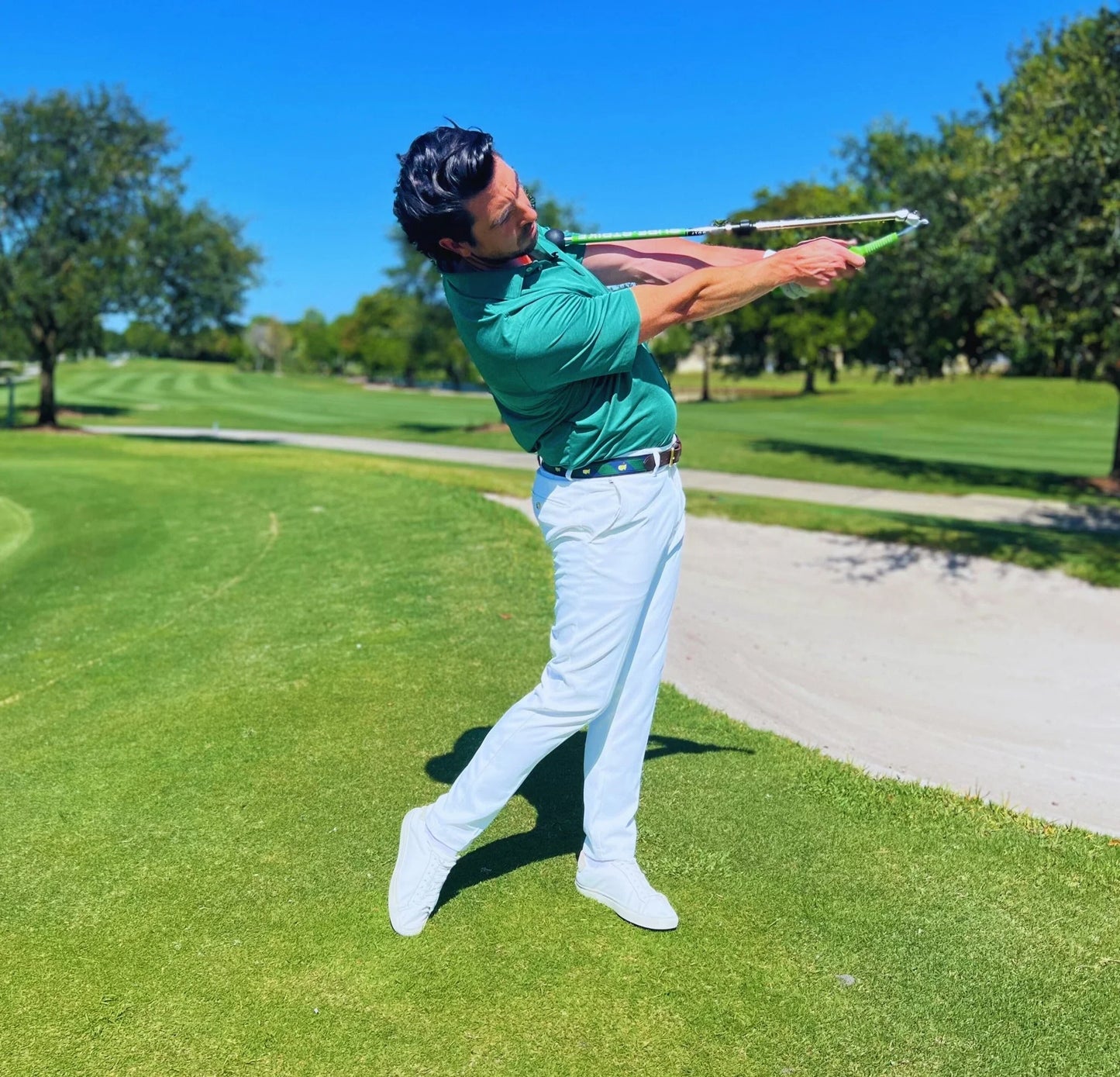 Golfer wearing green shirt and white pants practicing with Sure Strike golf training aid on green course