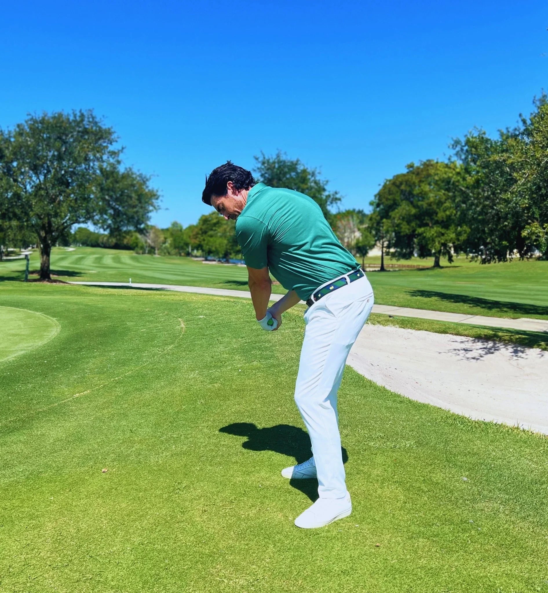 Golfer in green shirt and white pants practicing swing on sunny golf course near sand bunker