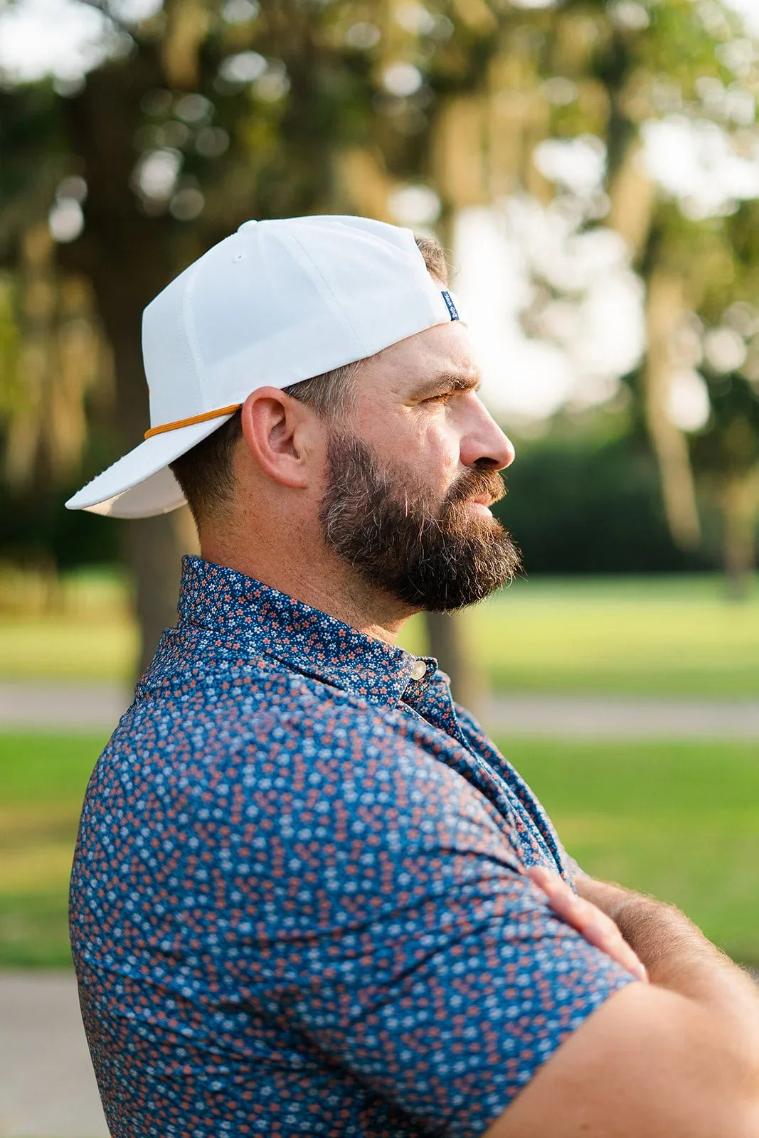 Man with beard wearing white cap backwards and blue floral shirt standing outdoors with arms crossed