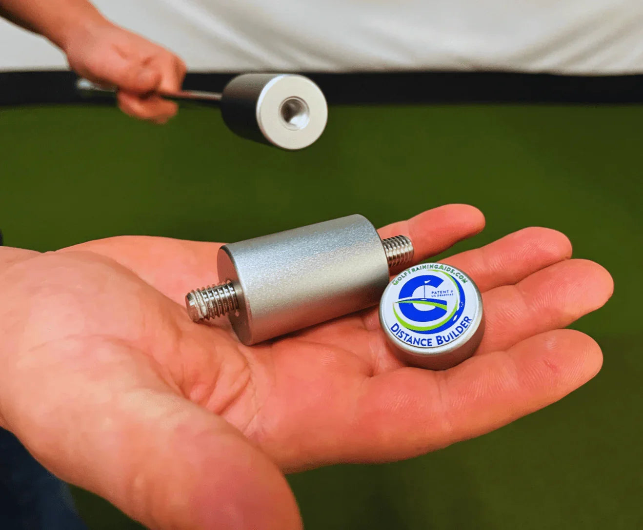 Hand holding a silver golf training aid labeled Distance Builder with green turf background