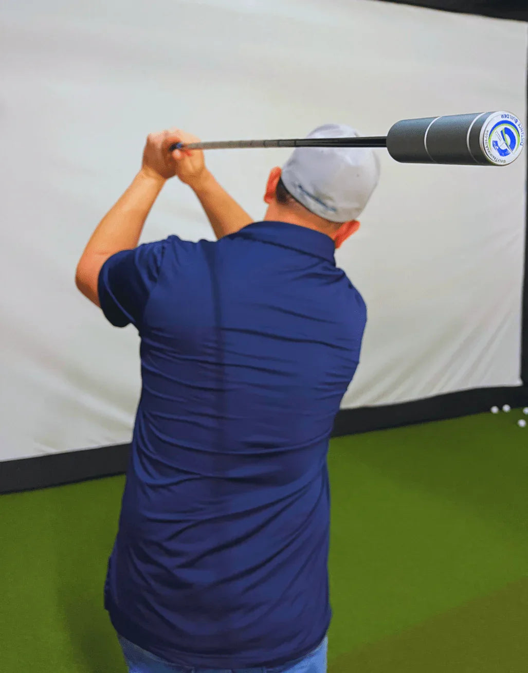 Man in blue shirt and gray cap swinging golf club with distance builder training aid on indoor putting green