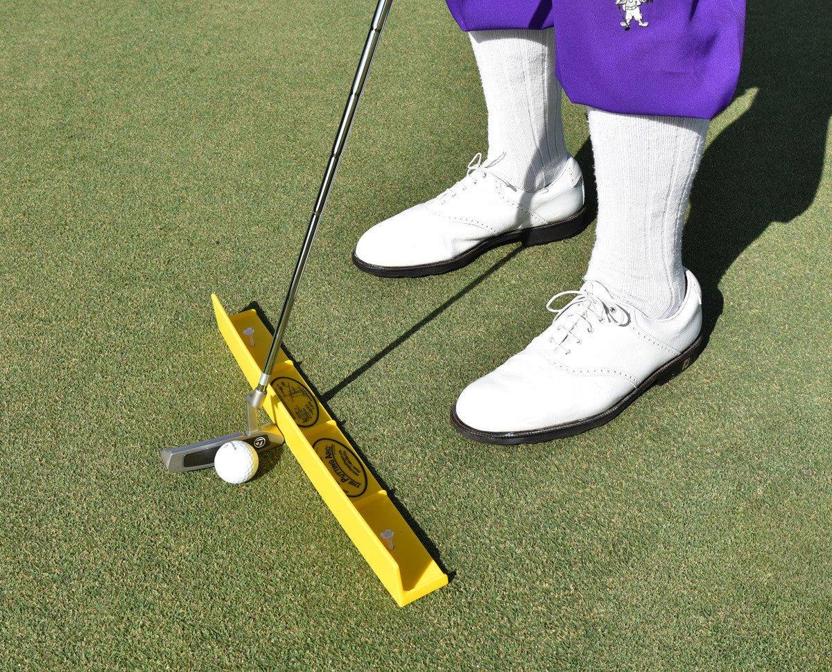 Golfer using a yellow putting arc training aid on green with white golf shoes and purple pants