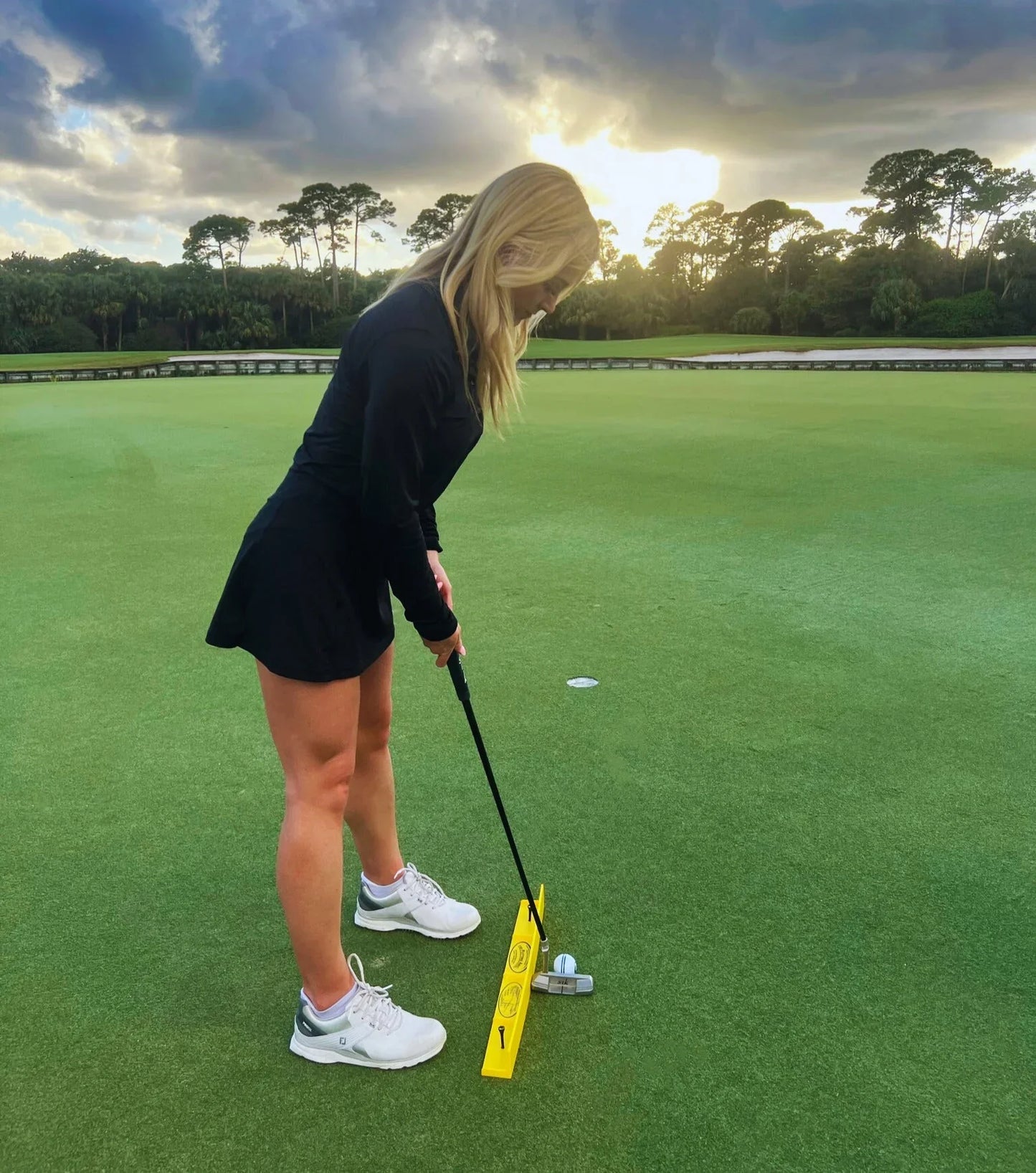 Woman in black dress practicing putting on golf green with Putting Arc training aid at sunset