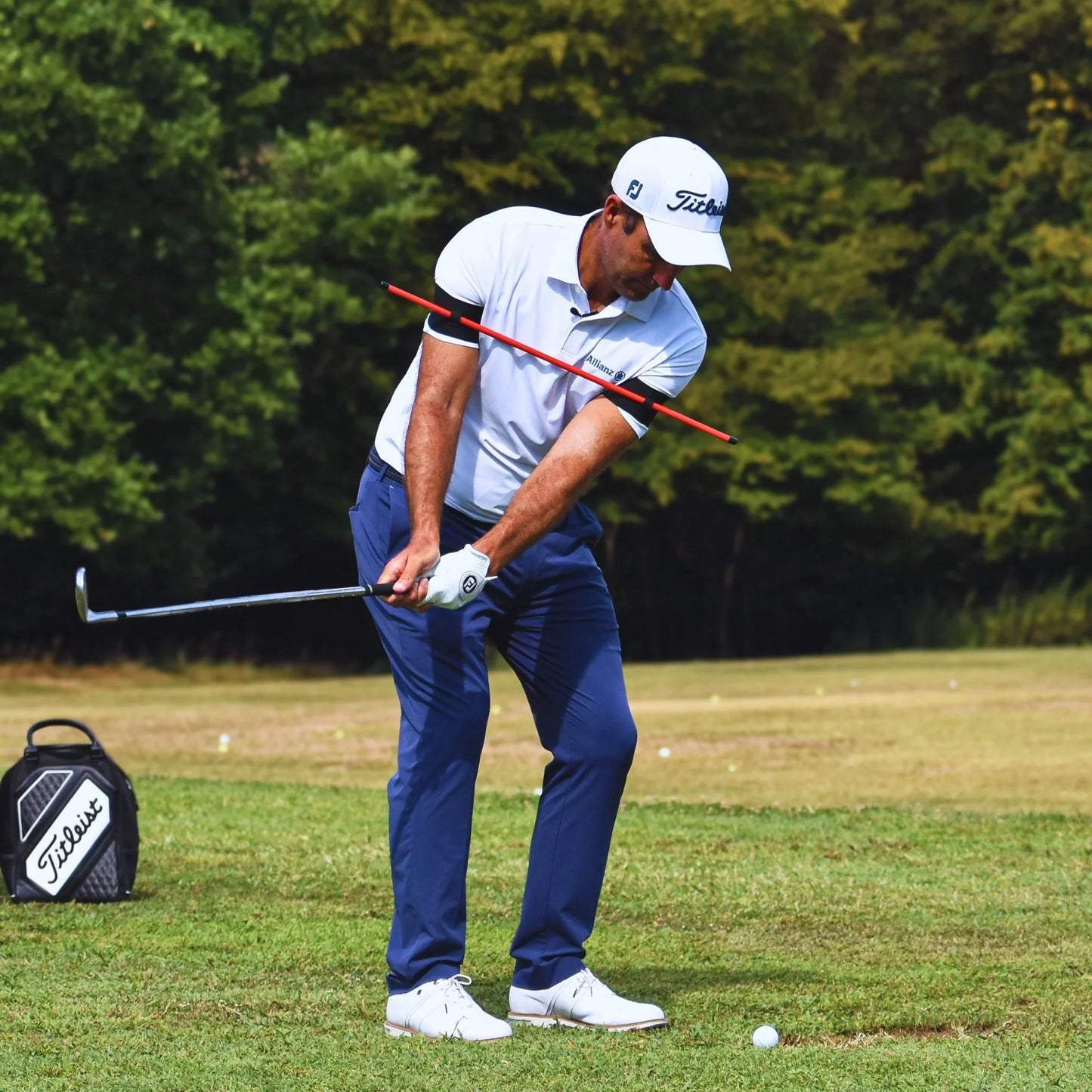 Golfer in Titleist cap and white shirt practicing swing on grassy golf course with trees background