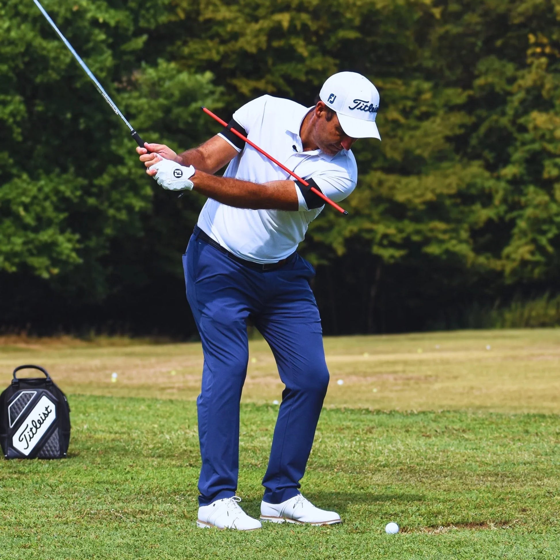 Golfer in white shirt and blue pants practicing swing on grass field with golf bag nearby