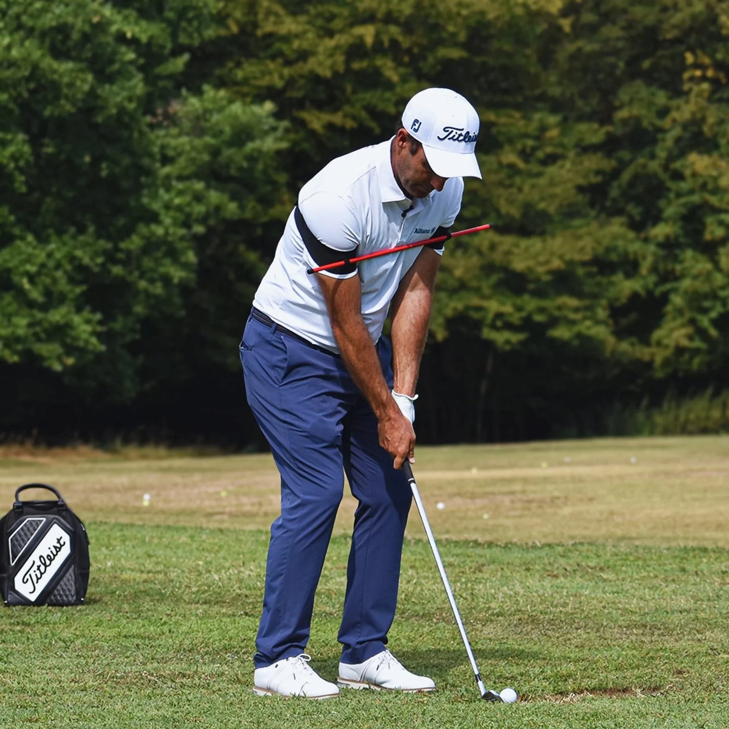 Golfer in white cap and polo using orange swing trainer on green golf course with Titleist bag