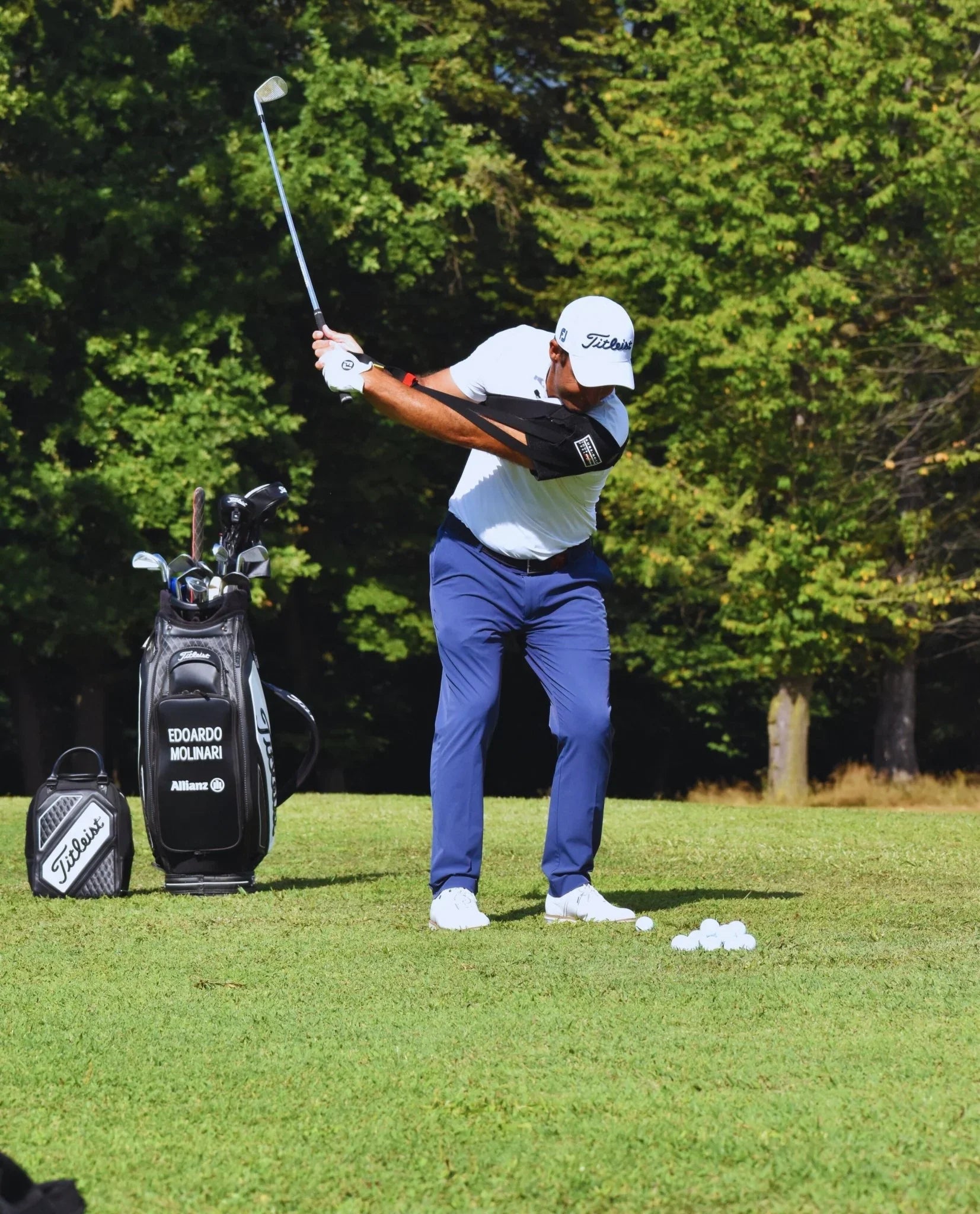 Golfer practicing swing with swing trainer on arm on grassy course with golf bag and balls