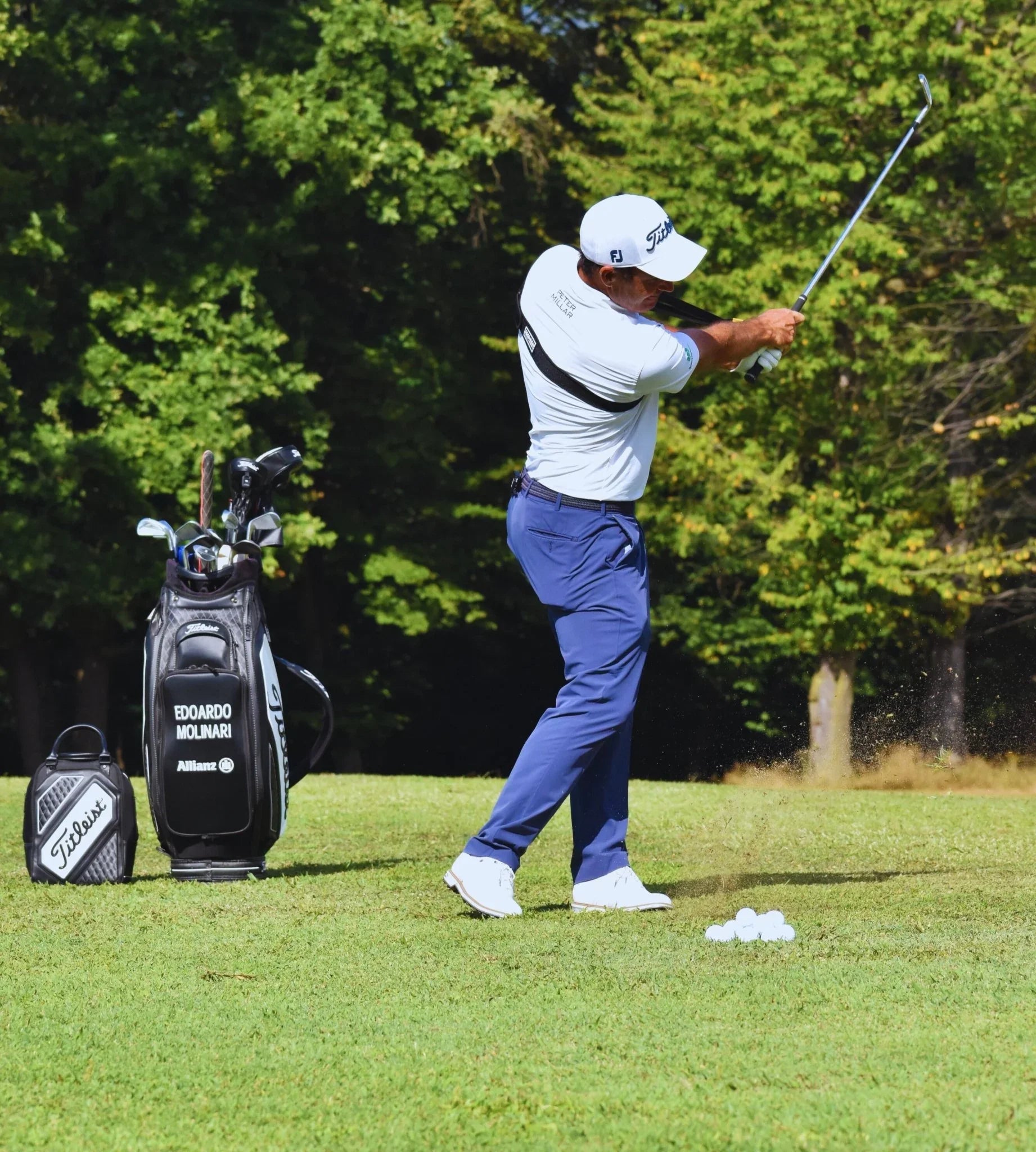 Golfer in blue pants and white shirt swinging club on green golf course with golf bag and balls nearby