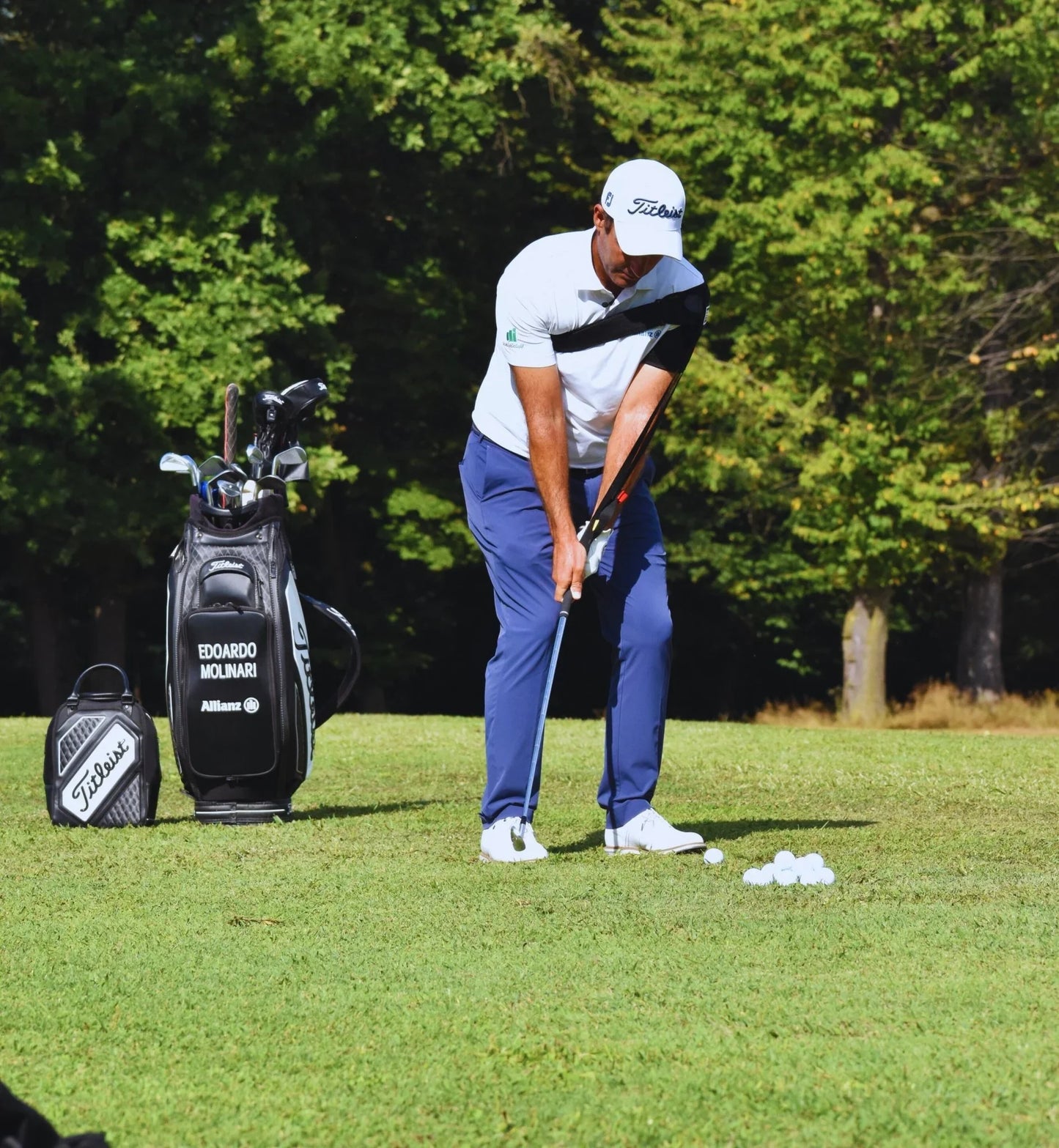 Golfer wearing white and blue practicing golf swing with Titleist clubs and bag on green field
