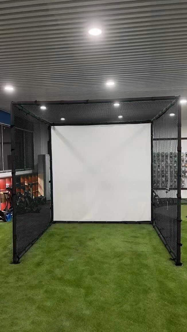 Indoor golf driving cage with black metal frame, protective netting, and white simulator impact screen on green turf